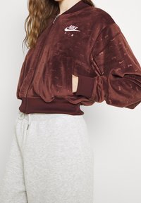 Burgundy sametová bomber bunda s žebrovanými manžetami a spodním lemem, s bílým logem Nike Air a decentními vzory loga. Nošena s světle šedými tepláky.