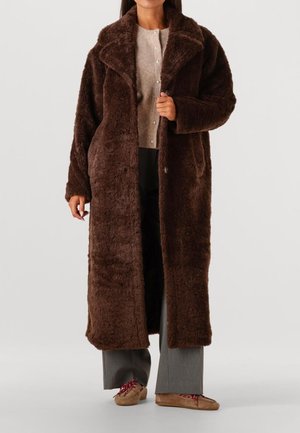 Bruine, fuzzy lange jas met een brede kraag, voorzien van een dubbelrijgsluiting en voorzakken, gelaagd over een licht sweater en grijze broek.