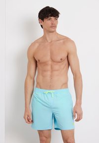 Pantaloni da bagno azzurri chiari realizzati in tessuto ad asciugatura rapida, con vita elastica munita di un laccio regolabile giallo neon e un piccolo dettaglio del logo.