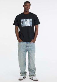 T-shirt en coton noir avec un imprimé graphique central gris. Assorti à un jean en denim clair et décontracté et des baskets blanches avec des accents noirs.