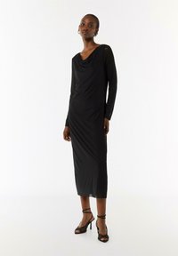 Robe noire à manches longues avec col drapé, en tissu lisse. Longueur genou avec fentes sur les côtés, associée à des sandales à talons hauts noires.