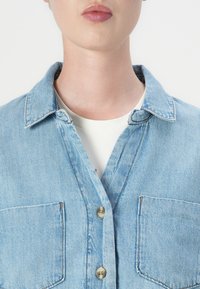 Camisa de ganga azul-claro com colarinho, dois bolsos no peito e botões em tom dourado. Apresenta uma textura ligeiramente desbotada e um corte relaxado.