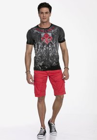 Grafisches T-Shirt in Schwarz und Grau mit rotem Fleur-de-Lis-Design, kombiniert mit roten Cargo-Shorts und schwarzen Sneakers mit weißen Sohlen.
