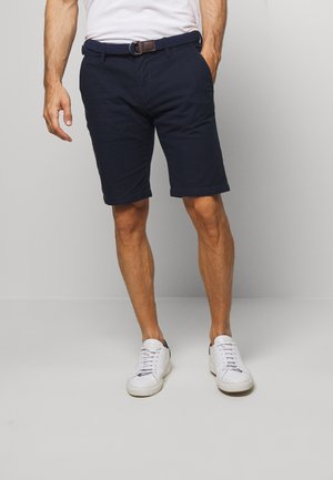 Shorts en coton bleu marine avec ourlets retroussés, dotés de poches latérales et d'une ceinture tressée. Portés avec des baskets blanches et un t-shirt blanc uni.