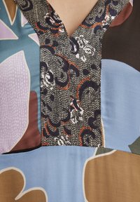 Mehrfarbiger Oberteil mit floralem und abstraktem Muster. Verfügt über einen V-Ausschnitt, glatte Texturen und eine Kombination aus blauen, lila, braunen und schwarzen Elementen.