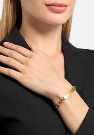 Brazalete de oro con un centro rectangular y plano, y un diseño de cadena enlazada. Lleva puesto en una muñeca, con un fondo de blaser negro.