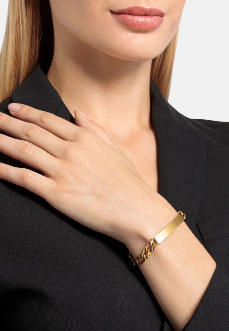 Brazalete de oro con un centro rectangular y plano, y un diseño de cadena enlazada. Lleva puesto en una muñeca, con un fondo de blaser negro.