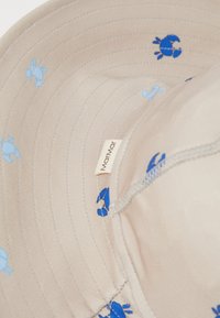 Beige bucket-hatt med blå krabbmönster. Insidan visar ett mjukt, texturerat tyg och en sydd etikett för varumärket.