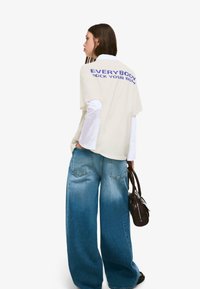 Beżowy, oversize t-shirt z niebieskim napisem "EVERYBODY ROCK YOUR BODY" na plecach, połączony z szerokimi, jasnoniebieskimi dżinsami i ciemną torebką.