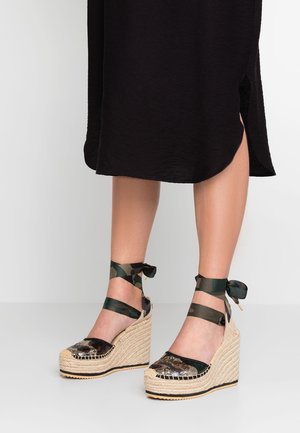 Wedge espadrilles with a woven jute sole, black and green patterned fabric upper, and a satin ribbon tie around the ankle.