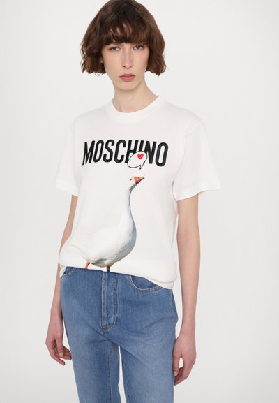 Balta medvilninė marškinėliai su žąsies grafika ir „MOSCHINO“ juodu tekstu; derinama su šviesiai mėlynomis džinsinėmis kelnėmis.