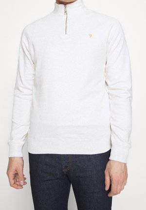 Hombre con suéter blanco de media cremallera con pequeño logo dorado en el pecho izquierdo y jeans azul oscuro, de pie contra un fondo liso.