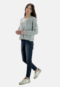 Lichtgrijze gebreide trui met losse mouwen, knoopsluiting, gecombineerd met donkerblauwe spijkerbroek en witte sneakers met beige accenten.