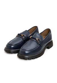 Mocassins en cuir bleu marine avec semelles noires épaisses et détail métal doré en forme de mors sur le dessus, inclinés côte à côte sur un fond blanc.