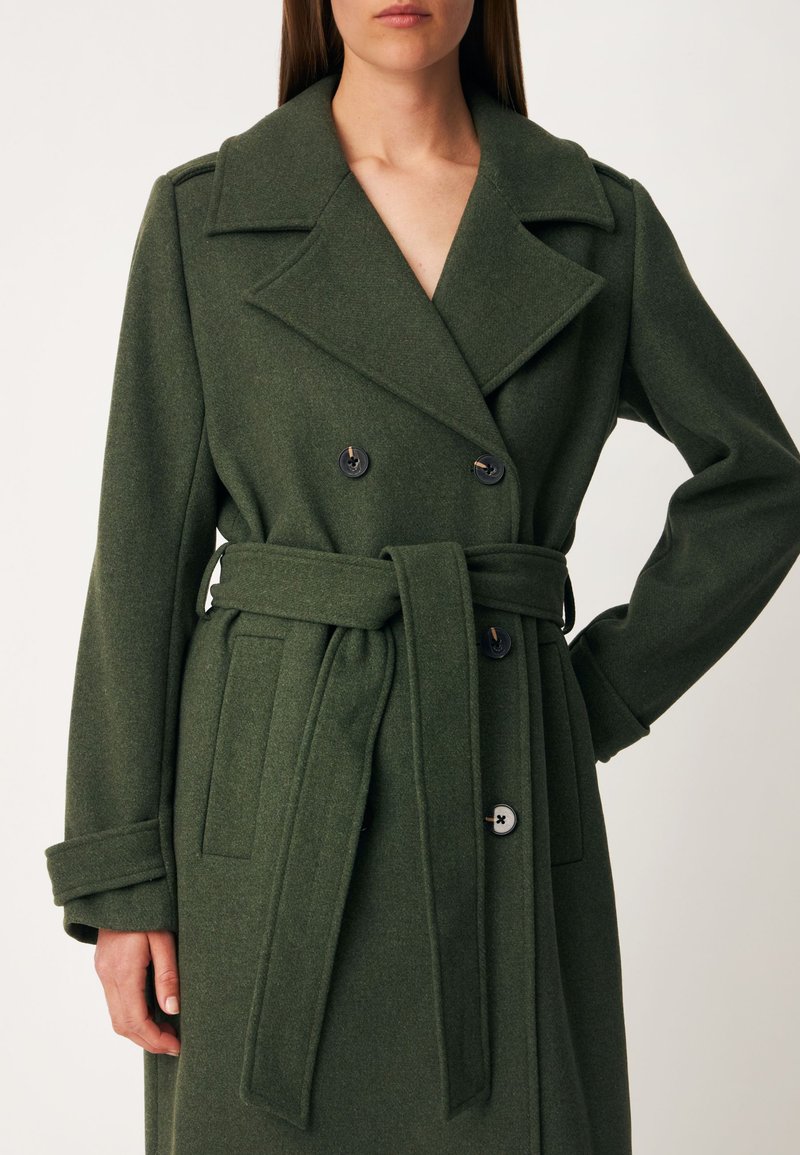 Femme portant un manteau en laine vert foncé à double boutonnage avec de larges revers et une ceinture nouée à la taille.