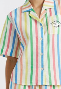 Maglietta da pigiama a righe in colori pastello—rosa, blu, verde, giallo—realizzata in un tessuto leggero, con taschino sul petto decorato da ricamo.