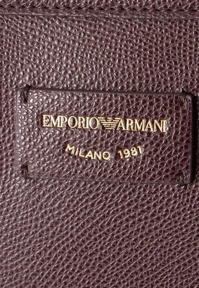 Κοντινό πλάνο καφέ δερματίνης με ραμμένη ετικέτα που γράφει "EMPORIO ARMANI MILANO 1981" με χρυσά γράμματα.