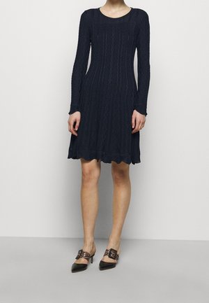 Robe longue en bleu marine à manches longues avec un motif losange texturé, un ourlet festonné et un col rond, associée à des escarpins noirs à bout pointu.