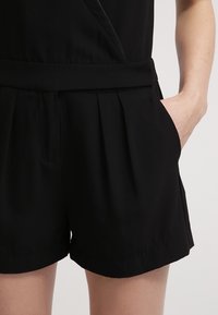 Svart rompers med figursydd överdel och plisserade shorts. Tillverkad av lättviktsmaterial, den har sidfickor och en mjuk textur.