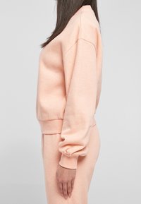 Sweat-shirt doux de couleur pêche en mélange de coton. Présente une coupe décontractée, des poignets côtelés et un col rond. Texture lisse dans l'ensemble.