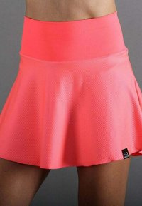 Falda atlética texturizada de un brillante color coral rosa con una cinturilla ancha, llevada por una persona de piel oscura frente a un fondo gris.