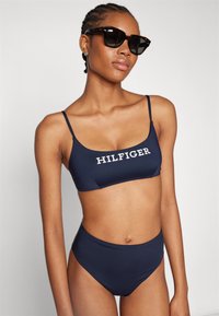 Jūrinei bikini komplektas su apvaliu iškirpimu, kuriame yra baltas "HILFIGER" logotipas, plonos petnešėlės ir juosmenį uždengiantis dugnas, pagamintas iš lygaus audinio.