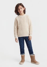 Kind mit mittellangen braunen Haaren, das einen beigen Strickpullover, hochgekrempelte dunkelblaue Jeans und beige Gummistiefel vor weißem Hintergrund trägt.
