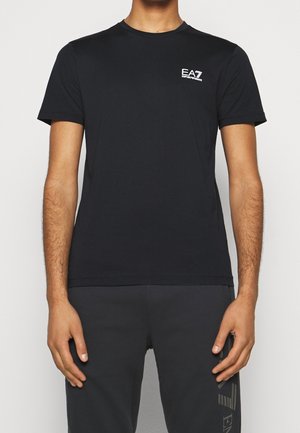 Mężczyzna noszący jednolitą czarną koszulkę z krótkim rękawem z małym białym logo EA7 Emporio Armani na lewej piersi oraz czarne spodnie z logo na lewym udzie.