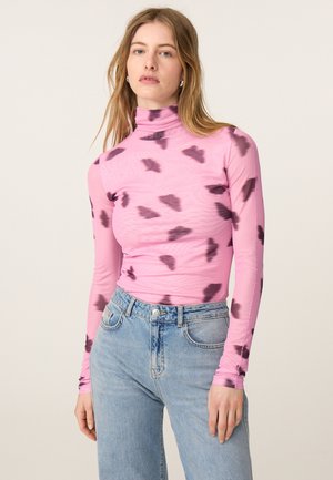 Vrouw met lang blond haar, die een roze coltrui draagt met donkerpaarse vlekken en lichtblauwe hooggetailleerde jeans tegen een effen achtergrond.