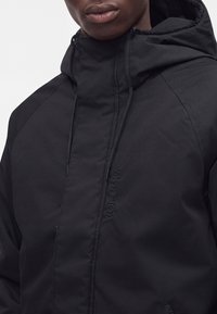 Personne portant une veste à capuche noire avec des cordons et ""Volcom"" brodé au centre devant.