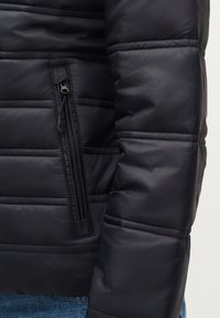 Μανίκι και πλαϊνή τσέπη με φερμουάρ μαύρου καπιτονέ μπουφάν, φορεμένα πάνω από μπλε τζιν, που δείχνουν το ράψιμο και την επενδυμένη υφή.