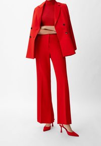 Blazer estructurado rojo con botones cruzados, combinado con pantalones de pierna ancha a juego y zapatos de tacón alto puntiagudos, todos con una textura brillante.