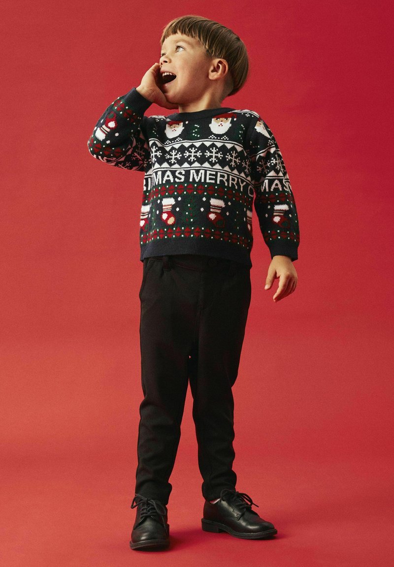 Bambino che indossa un maglione natalizio scuro con motivi festivi, pantaloni neri e scarpe nere, in piedi su uno sfondo rosso.