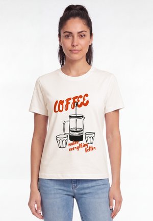 Vrouw met donker haar in een paardenstaart draagt een wit T-shirt met een illustratie van een French press en kopjes en de tekst "Coffee makes everything better."