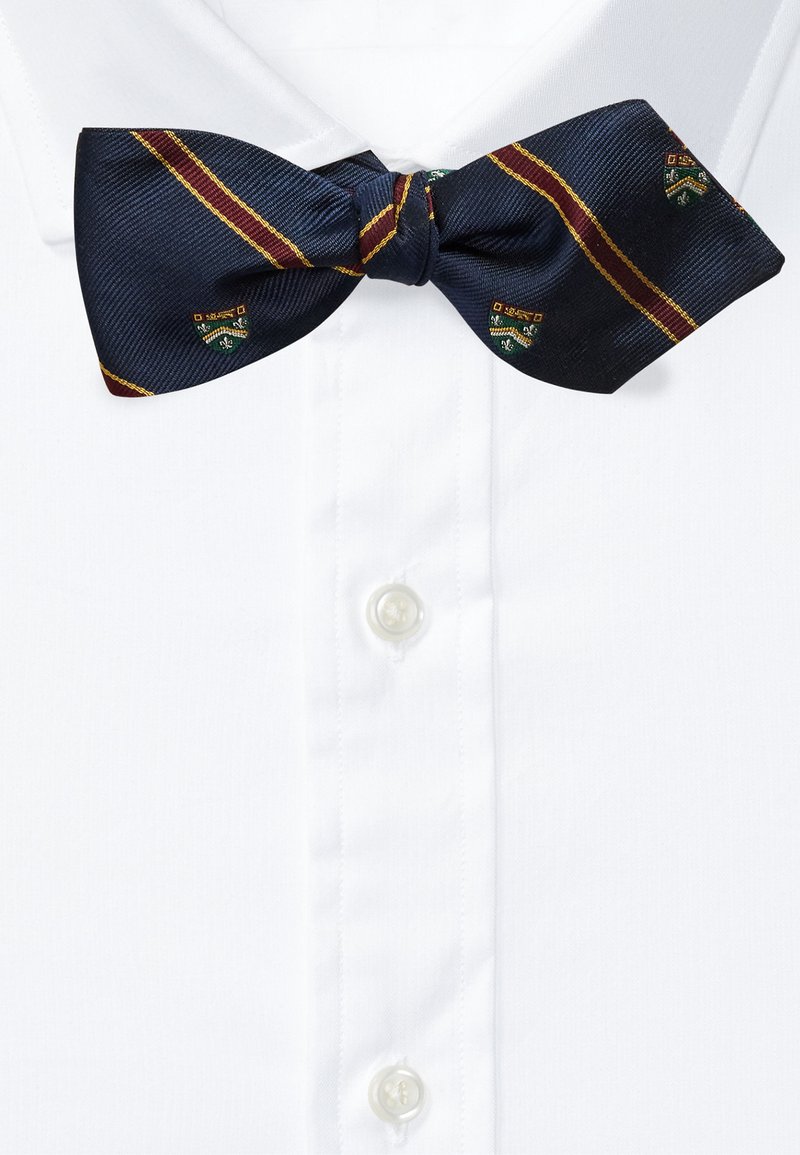 Navy silk bow tie with maroon and gold stripes and crested pattern, knotted at the center, positioned above a white button-down shirt.