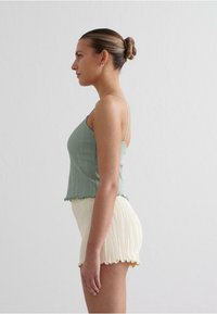 Top verde cu dungi și margine ondulată, asociază cu pantaloni scurți crem texturați cu un capăt volănat. Modelul este prezentat din profil.