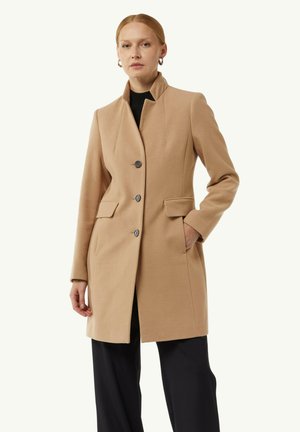 Beige wool coat with a high collar, two front pockets, and three buttons. Smooth texture, tailored fit, paired with black trousers.