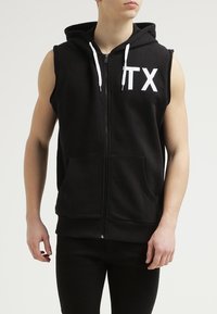 Felpa senza maniche nera con zip intera, cordini bianchi e la scritta "TX" in bianco sul lato sinistro. Materiale in cotone morbido, tasche frontali.
