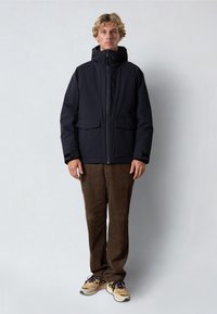 Svart isolerad jacka med huva, två framfickor och dragkedja framtill. Varm, mjuk textur. I kombination med bruna kordflöjlsbyxor och sneakers.