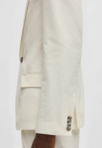 Giacca di costume beige chiaro in tessuto strutturato. Presenta una tasca sul petto, cinque bottoni metallici sui polsini e una vestibilità su misura.