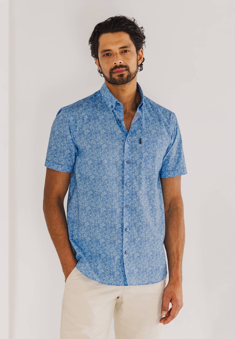 Blauw shirt met korte mouwen, voorzien van een bloemenpatroon, met knopen en een klein voorzakje, gecombineerd met lichtgekleurde broeken.