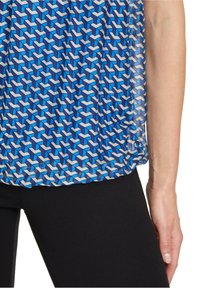 Modrý vzorovaný top s geometrickými tvary v odstínech modré a bílé, vyrobený z lehkého materiálu, doplněný černými legínami.