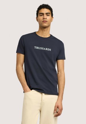 Giovane uomo che indossa una t-shirt Trussardi blu navy e pantaloni beige, in piedi con una mano in tasca davanti a uno sfondo semplice.
