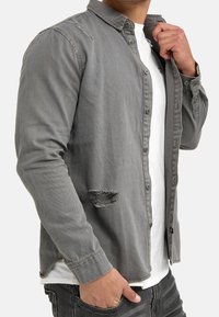 Grijs denim overhemd met een button-down kraag, voorzien van versleten accenten en een losse pasvorm, gedragen over een wit T-shirt.