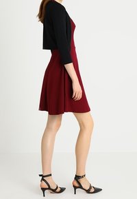 Vestido burdeos con falda acampanada, combinado con un cárdigan negro corto. Tacones negros de punta con correas en el tobillo y detalles de hardware mínimos.