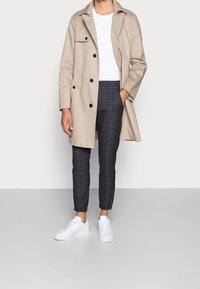 Beige trench coat med bruna knappar, vit T-shirt och mörka rutiga capribyxor. Vita sneakers kompletterar outfiten.
