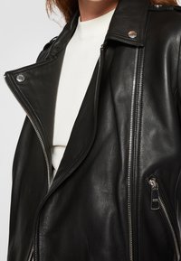 Černá kožená bunda se širokým límcem, asymetrickým zipem a texturovaným povrchem. Má stříbrné kovové doplňky a vrstvený design přes bílý top.