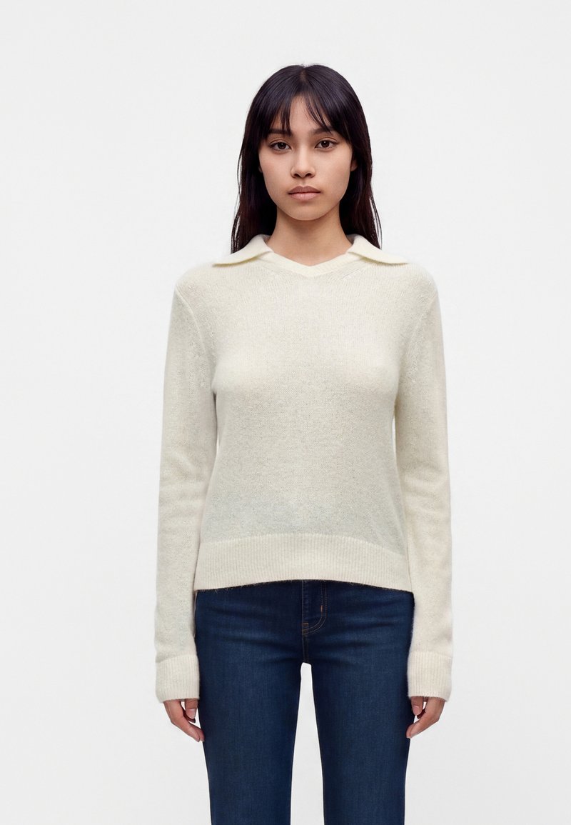 Jeune femme aux longs cheveux foncés portant un pull crème avec un petit col et un jean bleu foncé, debout devant un fond blanc uni.