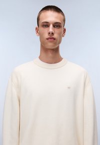 Maglione crema con dettagli a costine sulle spalle, scollatura rotonda e discreto logo. Texture liscia e vestibilità comoda.