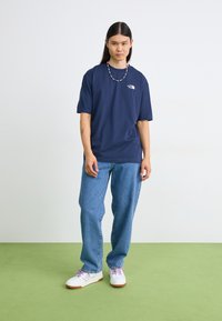 Mørkeblå t-skjorte med korte ermer og hvit logo, kombinert med lyseblå denimjeans og hvite sko med lilla aksenter. Grønn gulv bakgrunn.